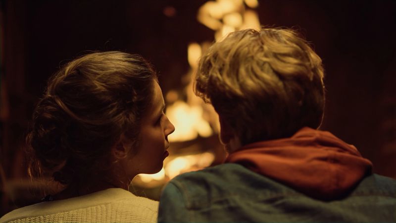Close up couple enjoy burning fire on date back view. Two lovers cuddle together by house fireplace. Loving boyfriend girlfriend looking eyes on romantic holiday evening. Vacation honeymoon concept.