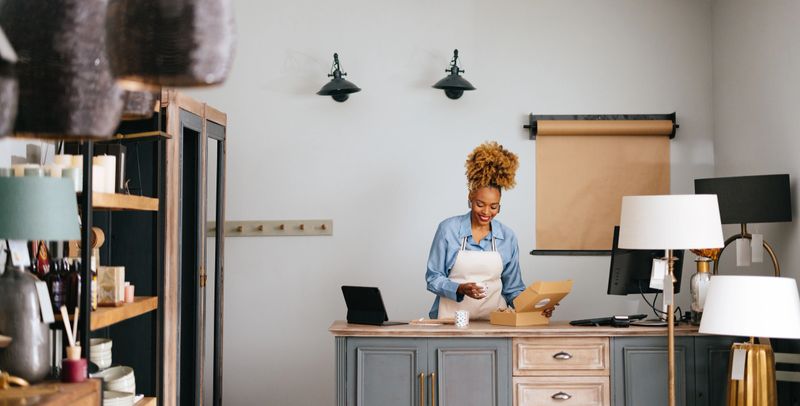 Smiling African-American woman preparing candles for delivery while standing at desk with computer, digital tablet and credit card reader.