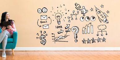 Woman sitting on teal chair holding tablet, looking at business doodles on wall.