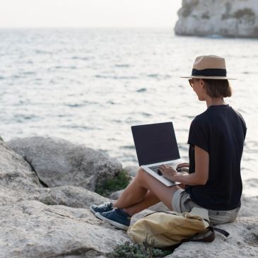 Person working on laptop by the sea on rocky shore, wearing hat and casual clothes.