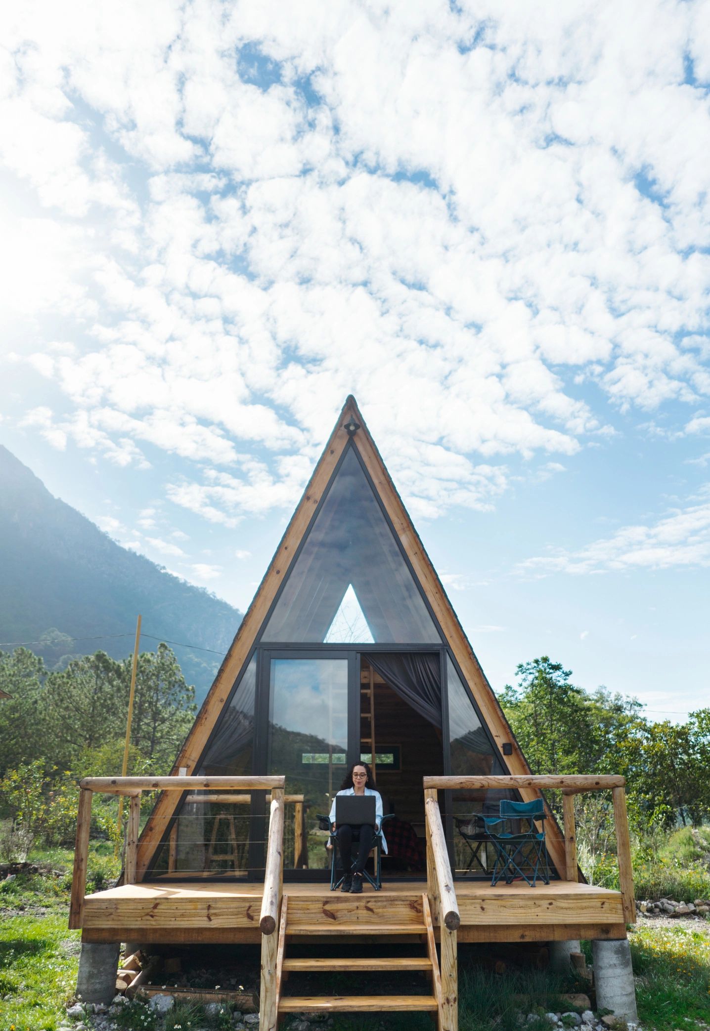 Woman working on laptop outside a wooden A-frame cabin in nature.