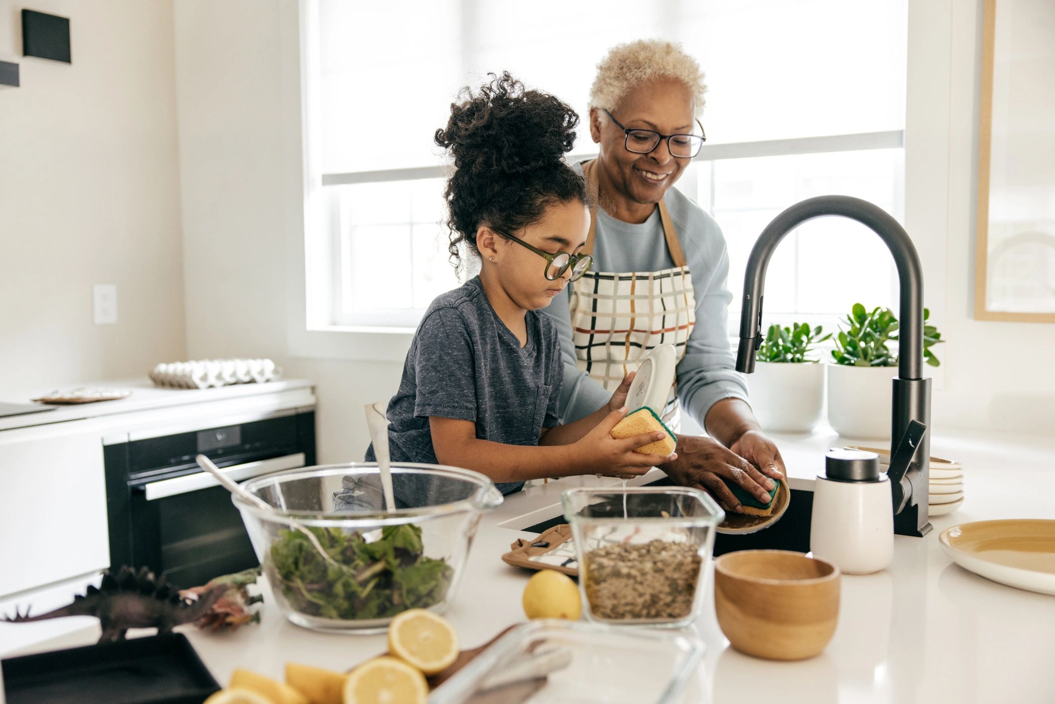 Grandmother and granddaughter washing dishes together in a bright kitchen.
