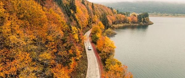 A car drives along a road beside a lake with vibrant autumn trees.