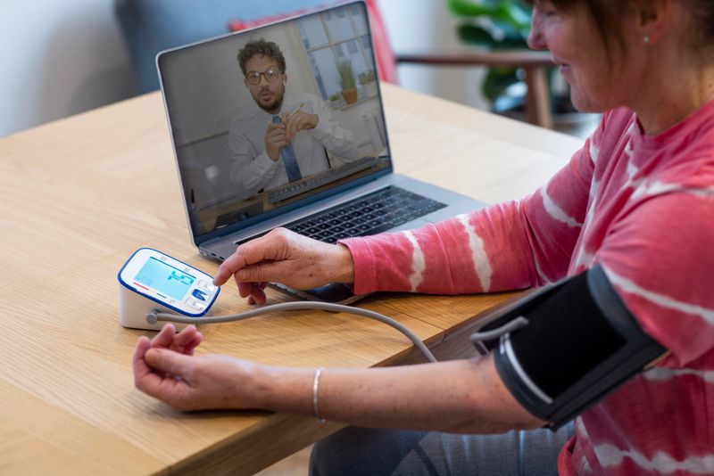Woman using a laptop to contact a medical professional about her health. She is siting at a table in a kitchen and is taking her blood pressure with a blood pressure gauge. She is in the North East of England.
