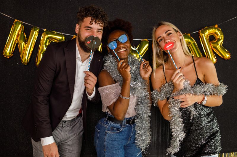 Front view of small group of diverse friends celebrating New Year's eve, dancing, having fun and smiling at camera while holding up photo booth props.