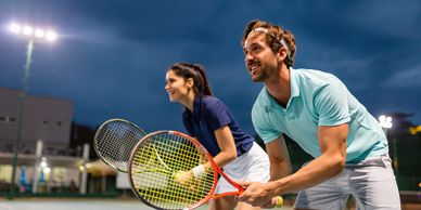 Two people ready to play tennis on an outdoor court at dusk.
