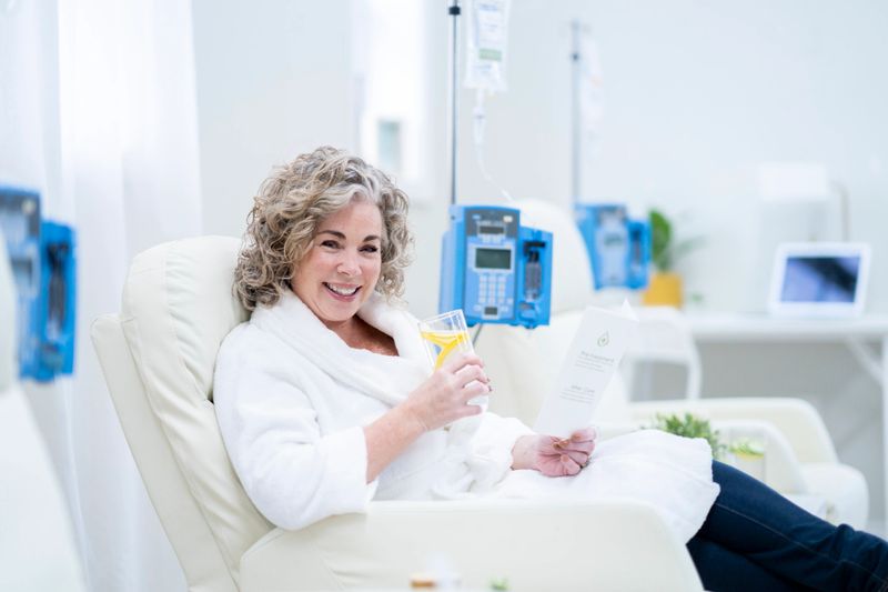 A beautiful middle aged woman sits in an oversized high back arm chair as she rests comfortably during her intravenous vitamin drip treatment.  She is dressed comfortably in a house coat and is sipping on lemon water ass he waits for the treatment to complete.