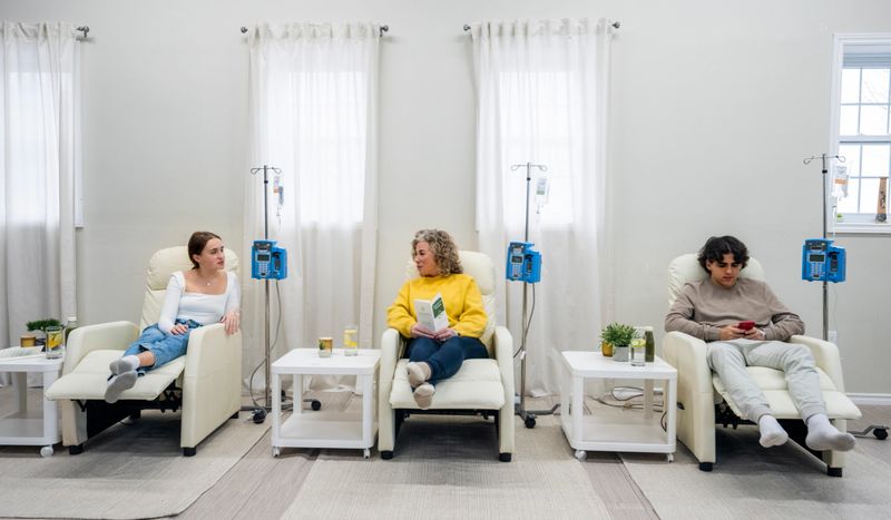 Three patients are seen in a Vitamin clinic as they sit in oversized high back chairs and receive their treatment intravenously.  They are each dressed comfortably as they talk among one another and keep occupied during the process.