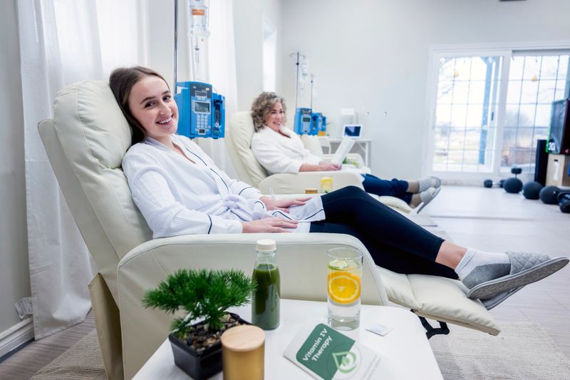 Two women are seen sitting in their individual oversized high back arm chairs as they wait for their intravenous vitamin drip to complete.  They are each dressed comfortably in warm housecoats and are sipping on lemon water as they share a conversation together.