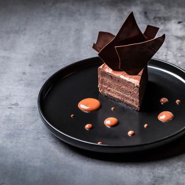 Elegant chocolate cake slice with decorative chocolate shards on black plate.