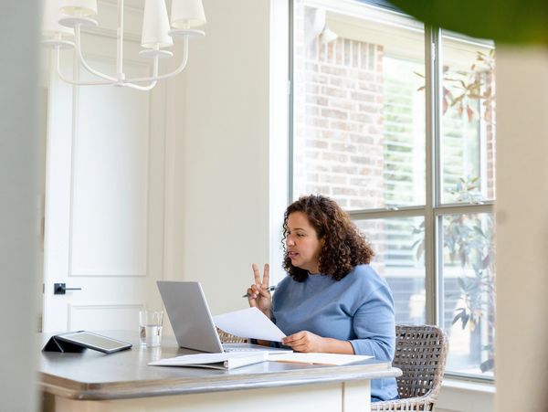 Woman in blue sweater gesturing peace sign during video call at home desk.