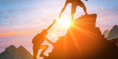 Two hikers silhouetted against a sunset, one helping the other climb a rocky peak.