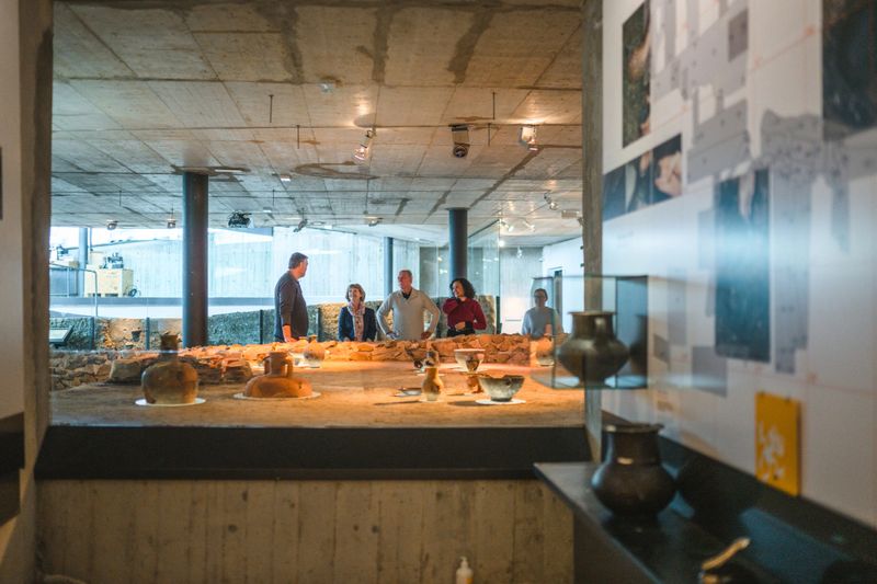 Shot over a large replica of Roman kitchen, showing a mixed group of older museum guests in distance, all listening to their male guide. Focus on the displayed objects.
