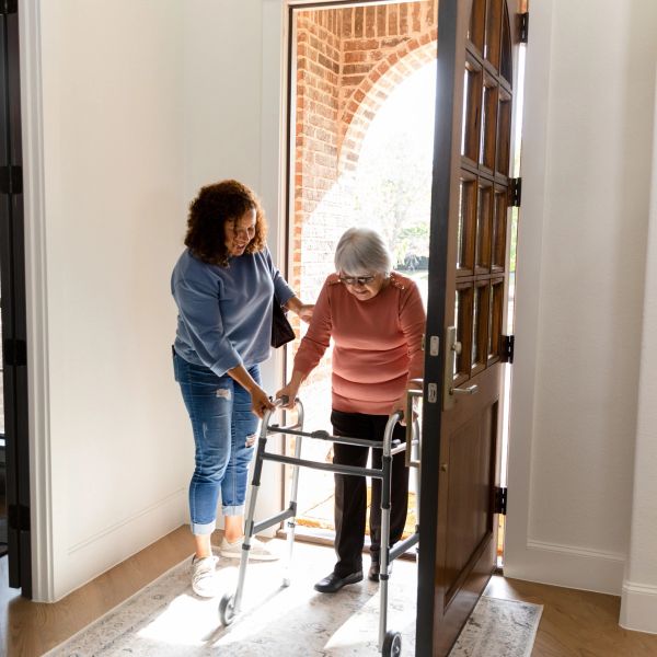 A woman helps an elderly lady with a walker enter a house through an open door.