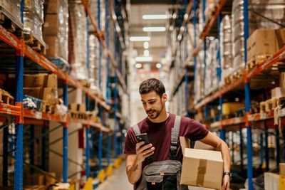 Warehouse worker checks phone while holding a box in a large storage area.