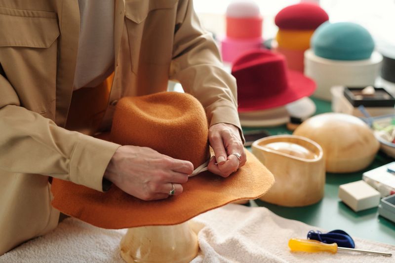 Close-up of young creative craftswoman fixing decorative shoelace arount new stylish felt hat of brown color and tying it while finishing work