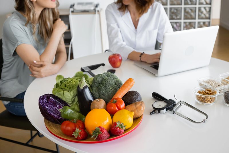 Cropped Dietitian typing on netbook and talking to content woman while looking at scream at desk with healthy food indoors