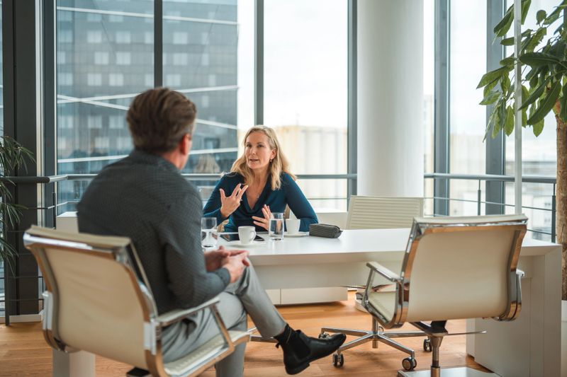Professional, mid adult Caucasian business woman sitting at an office desk with her back to the view of business district, interviewing a Caucasian male candidate, sitting with legs crossed,  only his back visible. Wide  shot. Focus on the woman.