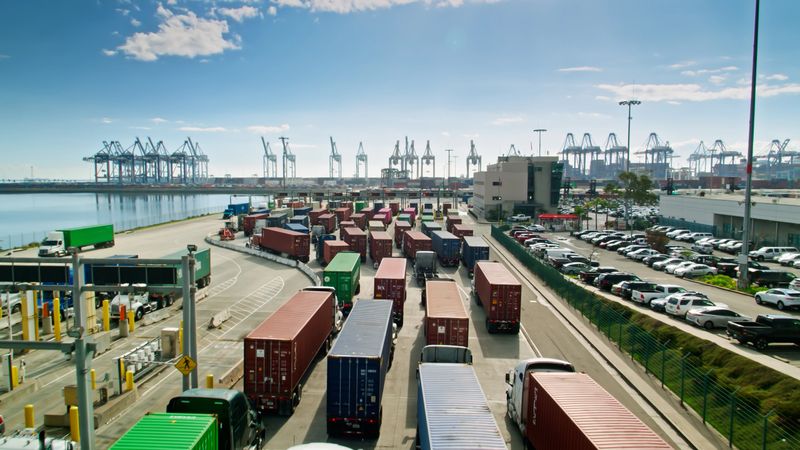 Drone shot of the front gate of a container terminal in the Port of Los Angeles on a sunny day. Trucks are lined up to leave and enter, and in the background massive container ships are sitting in their berths being loaded and unloaded by cranes.  

Authorization was obtained from the Los Angeles Harbor Department and Los Angeles Port Police for this operation in restricted airspace.
