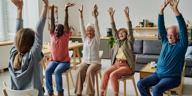 Group of seniors doing seated stretching exercises in a bright room.