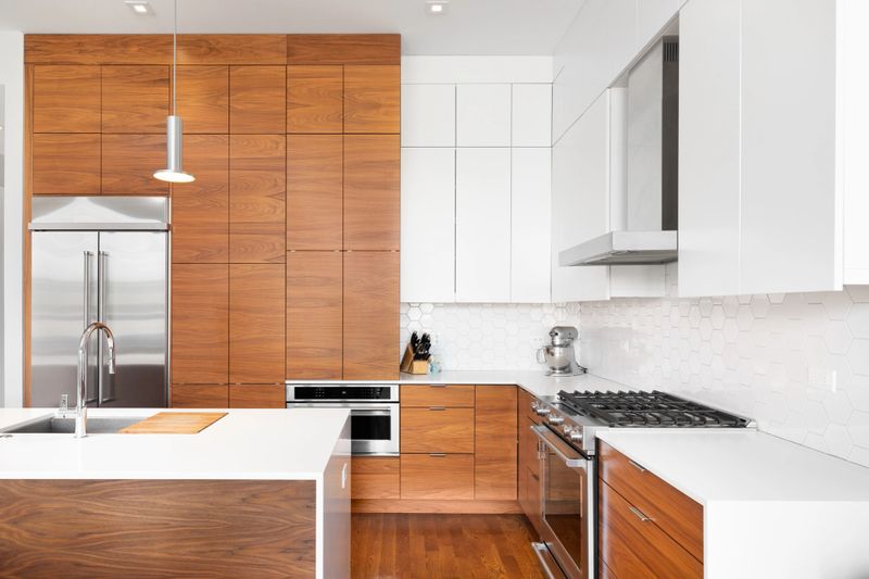 A modern, remodeled kitchen with wood and white cabinets, a white waterfall granite island, stainless steel appliances, and white pentagon backsplash tiles.