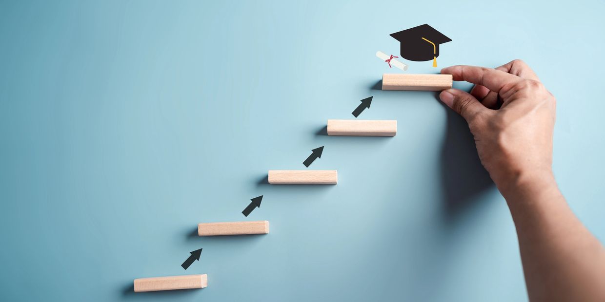 Hand placing a wooden block representing graduation as the final step in a staircase.