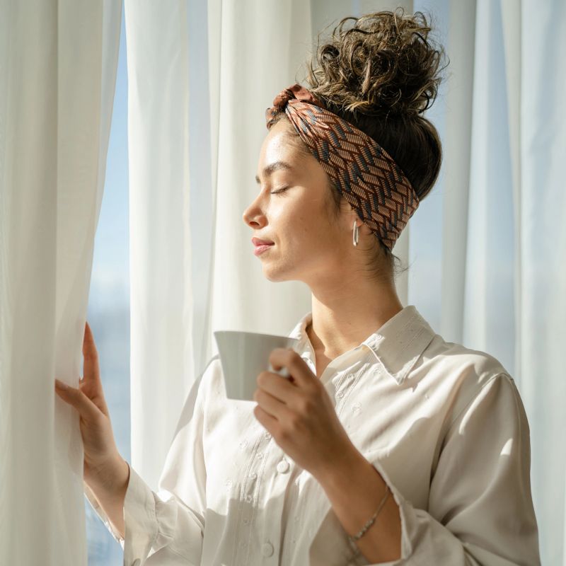One woman caucasian female standing at he window at home or hotel room in bright day holding a cup of coffee enjoy while taking a brake daily morning routine real people copy space wear white shirt