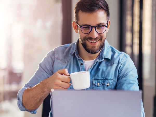 Man in glasses enjoying coffee while working on a laptop.