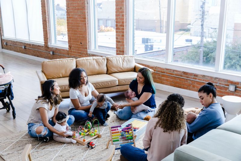 The diverse group of young mothers sit together and visit as their children play.