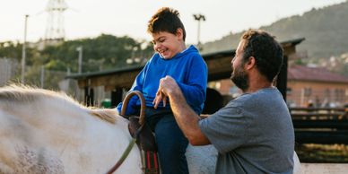 A boy joyfully rides a white horse with an adult assisting him.