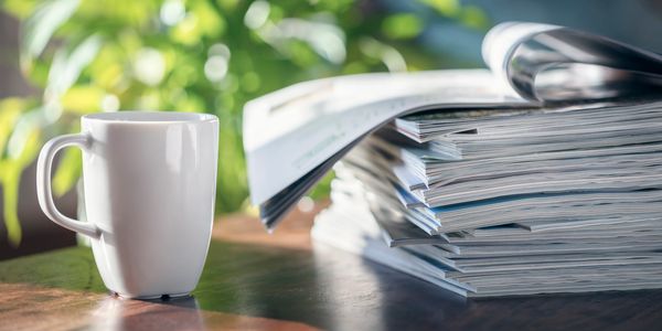 Cup of coffee with a stack of magazines placed on a wooden desk.