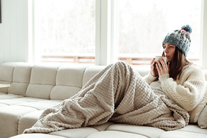 Beautiful young woman drinking hot tea wrapped in blanket on sofa at home.