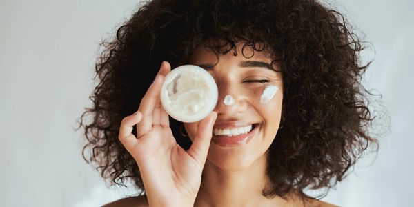 A smiling woman with curly hair holds a jar of cream, applying it to her face.