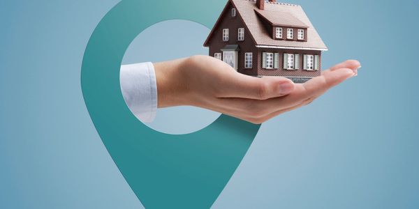 Hand holding a miniature house with a location pin background.