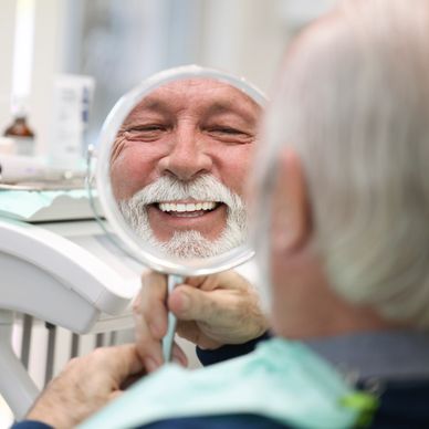 Elderly man happily admiring his teeth in a dental mirror.