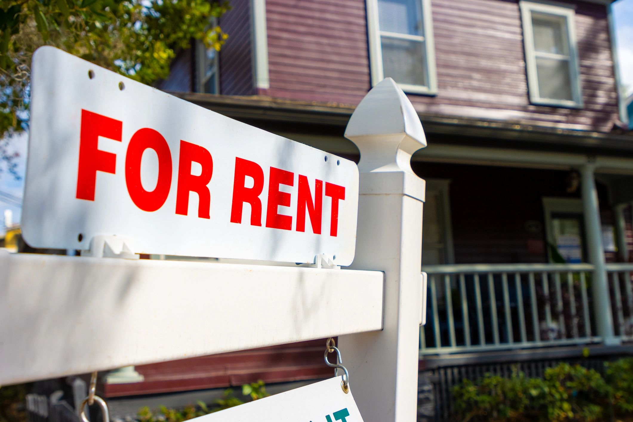 A white sign with red letters saying 'FOR RENT' in front of a house.
