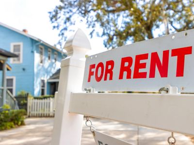 A 'For Rent' sign in front of a blue house on a sunny day.