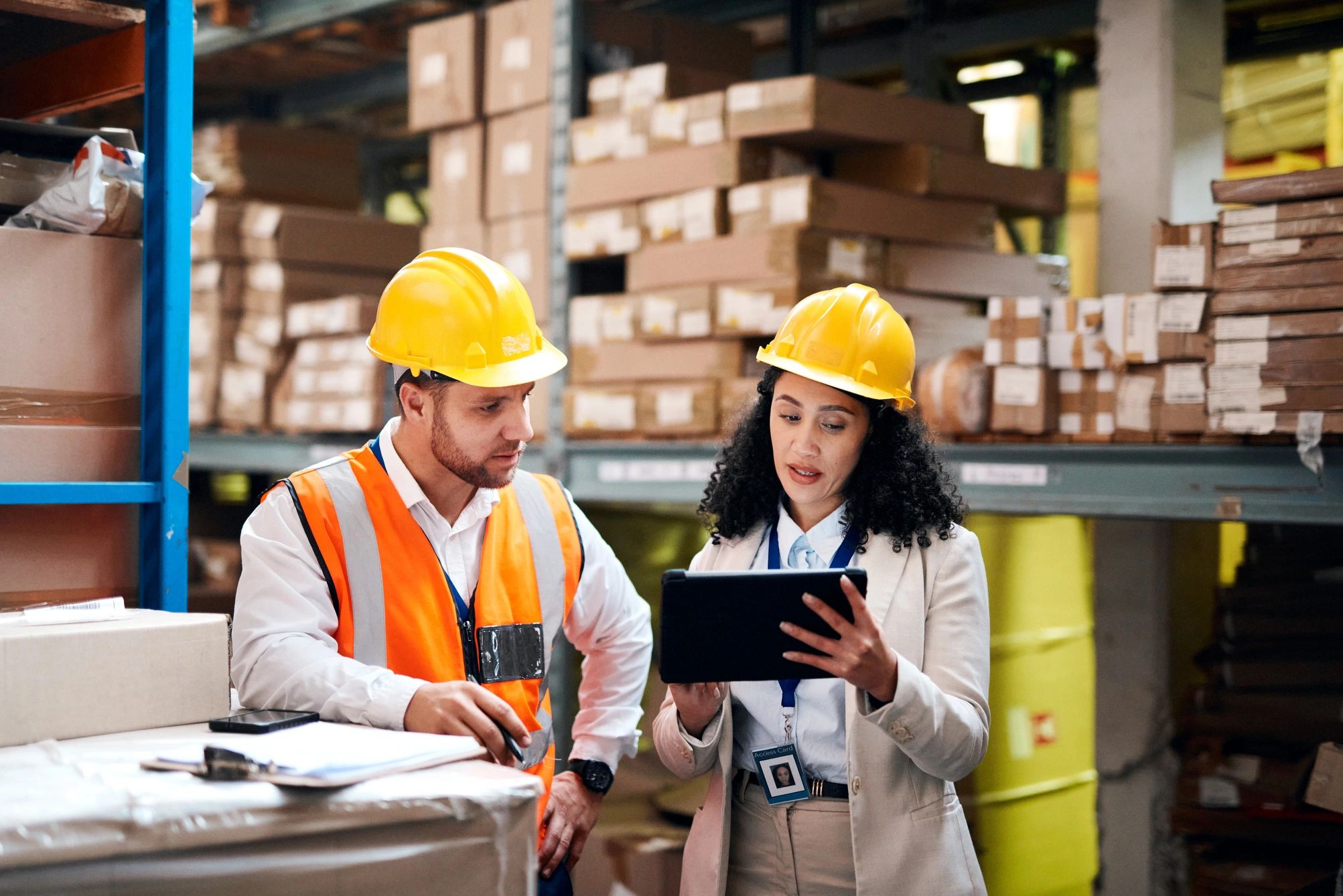 Two warehouse workers in hard hats discussing something on a tablet.