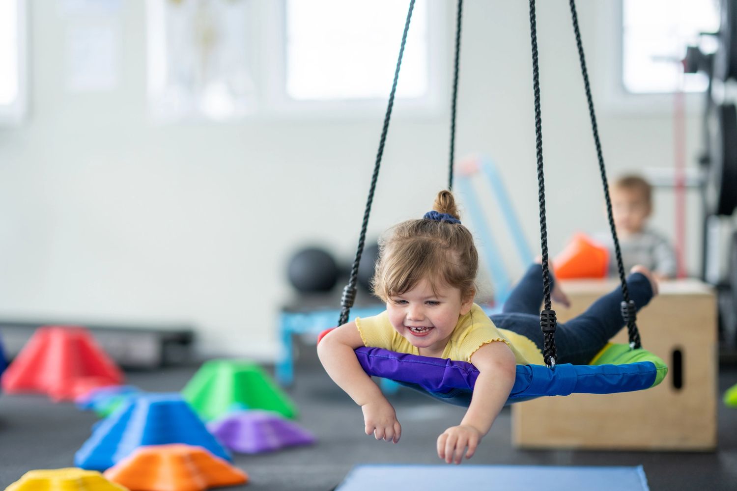 A young girl joyfully swings on a colorful suspended platform indoors.