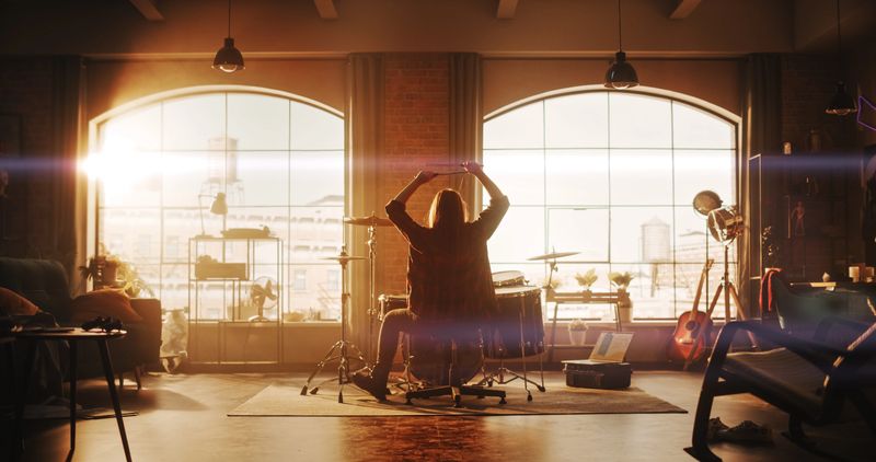 Person Playing Drums During a Band Rehearsal in a Loft Studio with Sunlight at Daytime. Drummer Practising Alone Before a Live Concert on Stage. Warm Color Editing.