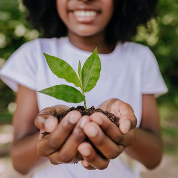 A smiling person holding a small green plant with soil in their hands outdoors.