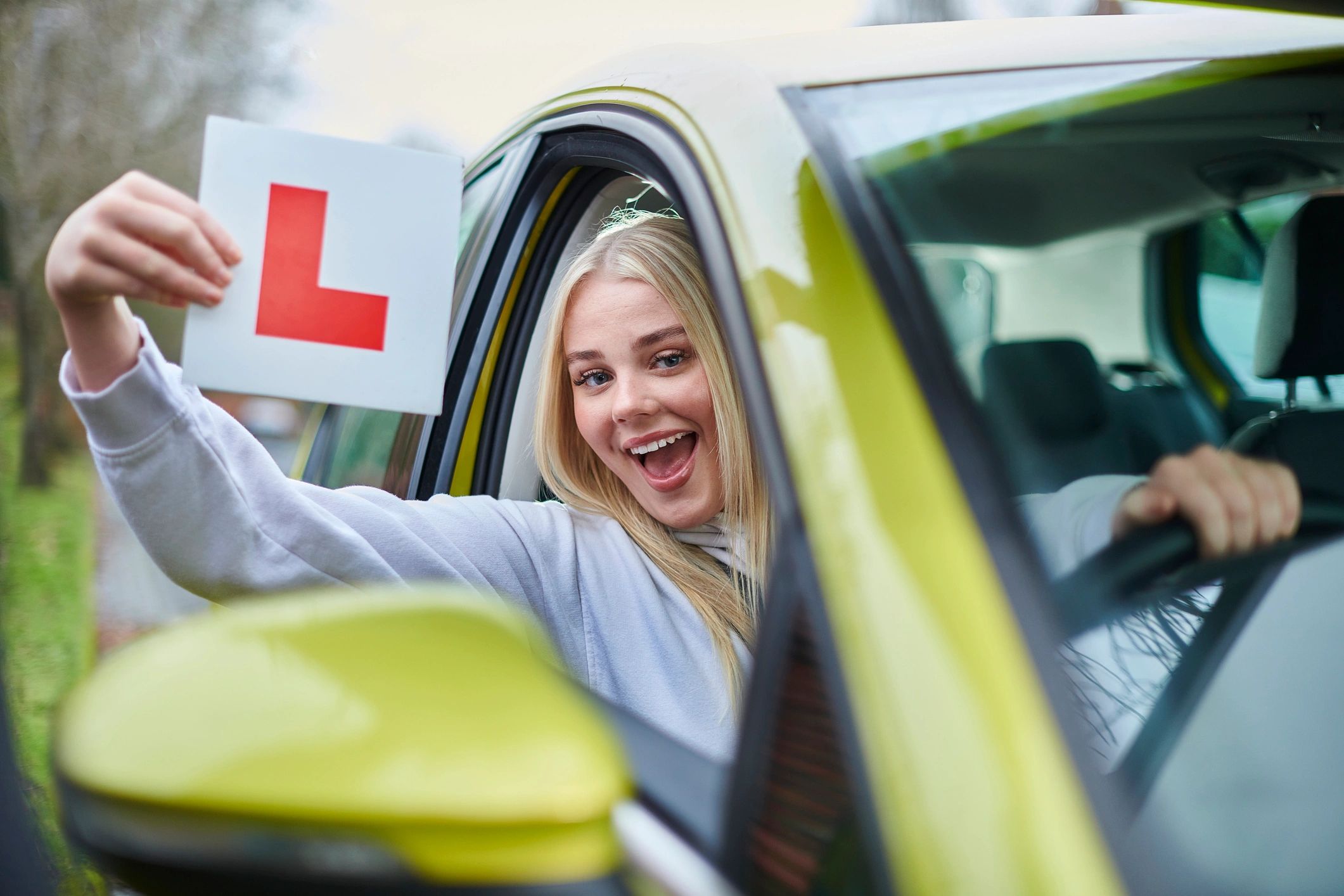 Happy young woman holding a learner driver L plate from car window.