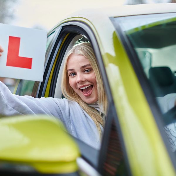 Happy young woman holding a learner driver L plate from car window.