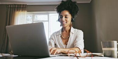 Woman working on a laptop, smiling with headphones in a cozy room.