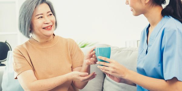 A nurse hands a blue cup to a smiling elderly woman on a sofa.
