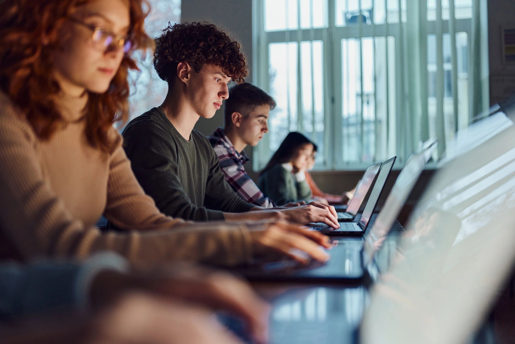 A group of young adults is seated in a classroom or computer lab, each focused on their laptops. 