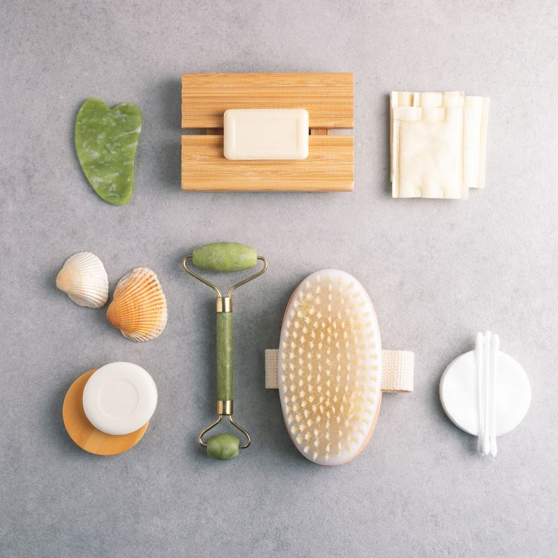 Self care products arranged on grey stone background, square composition. Dry massage brush, cotton pads, soap and jade stone gua sha massagers.