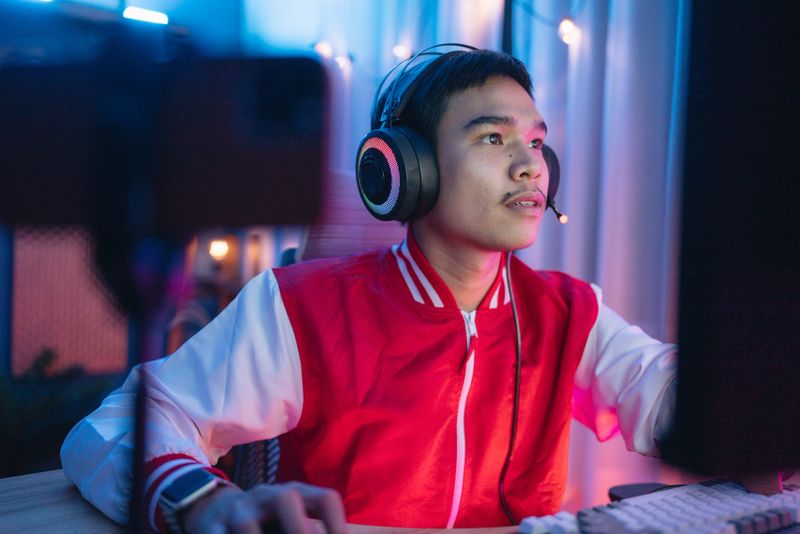 Asian man in headphones making live stream on smartphone while playing fps game on pc at home