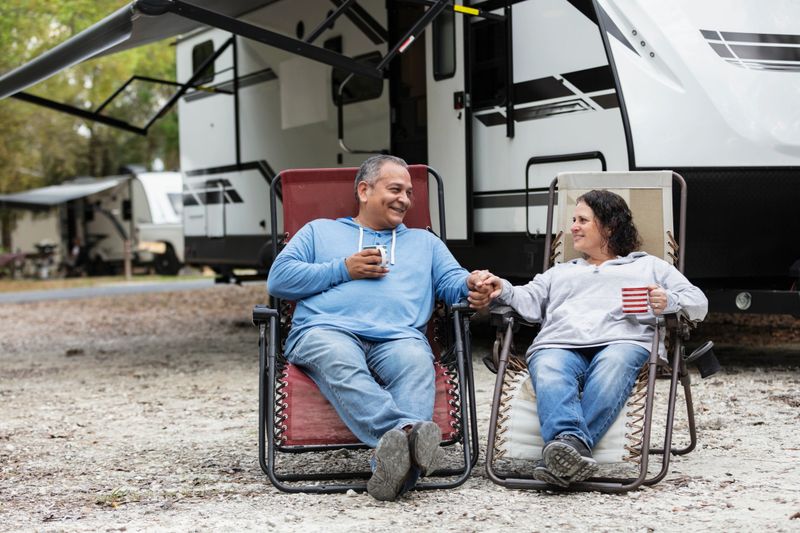 A mature multiracial couple sitting together in chairs by a camper trailer in an RV park, drinking coffee. He is Hispanic and she is Middle Eastern and Caucasian. They are conversing, smiling, looking at each other, and holding hands.