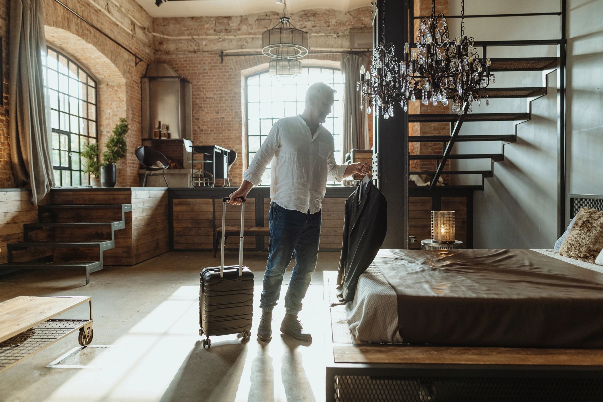 Man holding a suitcase and jacket in a stylish, sunlit loft bedroom.